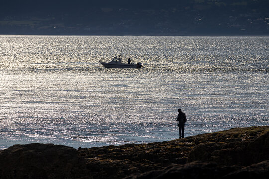 Silhouetted angler fishing from rocky shore with boat on shimmering water at Penmon Point on the Isle of Anglesey