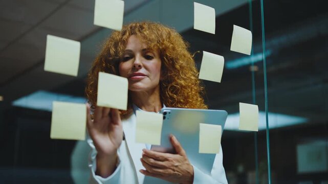 Confident redhead businesswoman in a modern office writing ideas on sticky notes attached to a glass wall, organizing a project