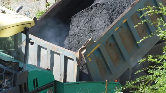 Heavy dump truck unloads hot steaming asphalt into the hopper of a paving machine during road construction works. 