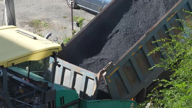 A dump truck is unloading hot asphalt mixture into the hopper of an asphalt paver during road construction works. Heavy machinery performs laying of a new asphalt pavement on the street.