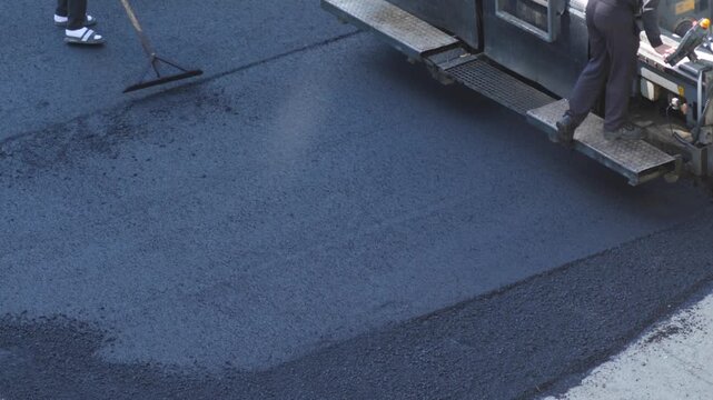 An asphalt paver evenly distributes hot new asphalt pavement on a city road. A construction worker manually levels the edge of the seam with a hand tool during road paving operations