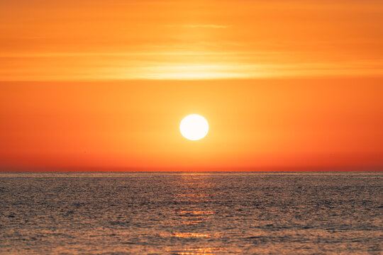 Bright glowing sun rising over calm ocean horizon with water reflections under orange sky