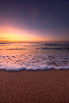 Vertical view of gentle ocean wave foam washing onto sandy beach under purple and orange sunset sky
