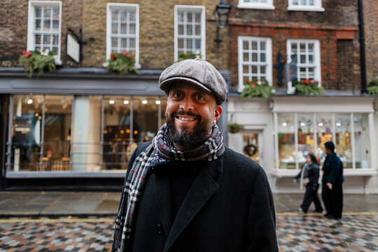 A smiling male wears a flat cap and scarf on a winter city street
