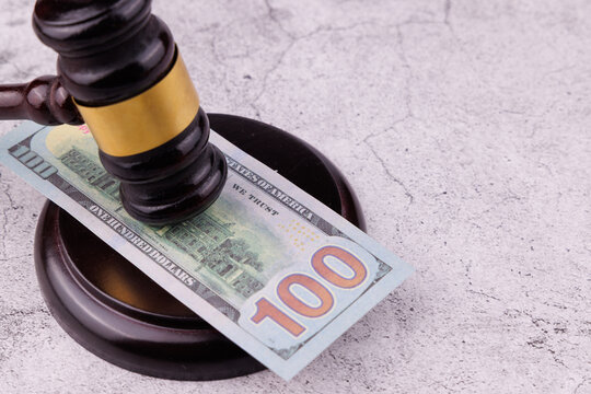 Straight eye level view of a dark mahogany judge gavel on top of hundred Dollar banknotes resting on a wooden sound block on a gray concrete tabletop, financial law enforcement