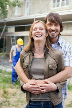 Young couple at site embracing, construction worker in background