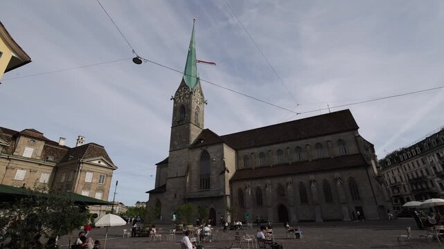 View of the historic Fraumunster church with its iconic green spire and clock tower, situated in a bustling square with people at outdoor tables in Zurich, Zurich, Switzerland.