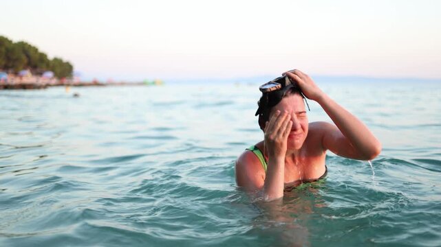 Woman removes diving mask and rubs her irritated eye after seawater gets inside while swimming causing burning discomfort and temporary vision trouble during seaside swim