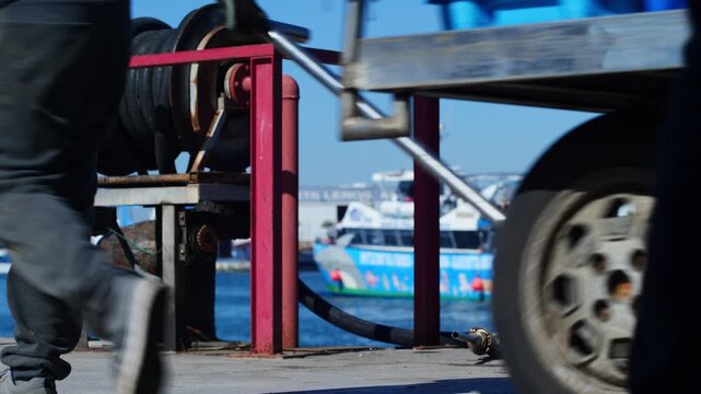 Close-up of a harbor gas hose with a traditional tabarquera boat blurred in the background and fishermen pushing a crate cart. Authentic maritime harbor scene. Santa Pola, Spain.