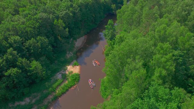 Friends kayaking features several colorful inflatable boats moving in separated groups through a narrow channel as the high drone tracks their route between dense trees small sandbars and shadow