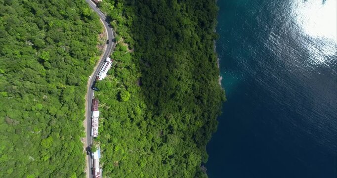 Winding coastal road cuts through rainforest beside dark blue sea in a vertical drone view with sharp bends tin roof roadside buildings rocky shoreline reflections and a few small vehicles below