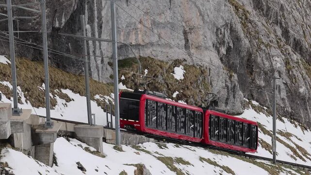 View of the red Pilatus Railway train ascending the steep, snowy track along the rocky cliffside of Mount Pilatus in Alpnach, Obwalden, Switzerland.