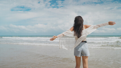 A young woman seen from behind stands on a sandy beach with open arms, enjoying the sea breeze under a bright blue sky © M Stocker