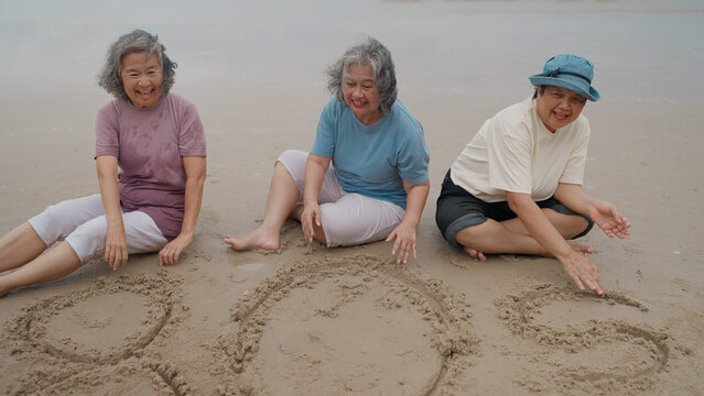 Three senior Asian women sit on the beach sand, smiling happily as they write "80s" together in the wet sand by the sea