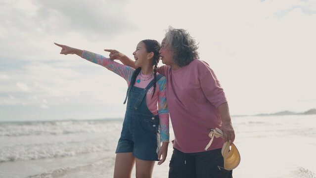 An Asian grandmother and her young granddaughter stand together on the beach, smiling as they point out into the distance towards the ocean