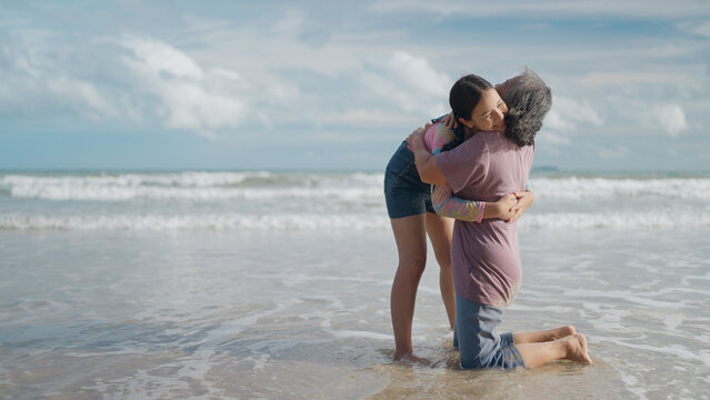 An Asian grandmother kneels in the shallow shore water and tightly hugs her young granddaughter, both smiling warmly against the gentle ocean waves