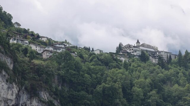 View of residential houses nestled on a lush green hillside with rocky cliffs and cloudy sky in Lucerne, Lucerne, Switzerland.