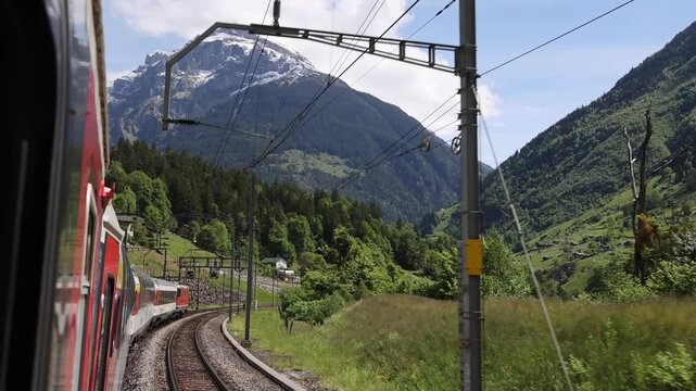 View of Gotthard Panorama Express SBB train traveling through the scenic mountains with lush green forests and snow-capped peaks in Lugano, Ticino, Switzerland.