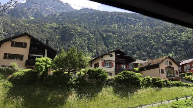 View of the Gotthard Panorama Express SBB route passing by traditional alpine houses and lush green hills with a dramatic mountain backdrop in Lugano, Ticino, Switzerland.