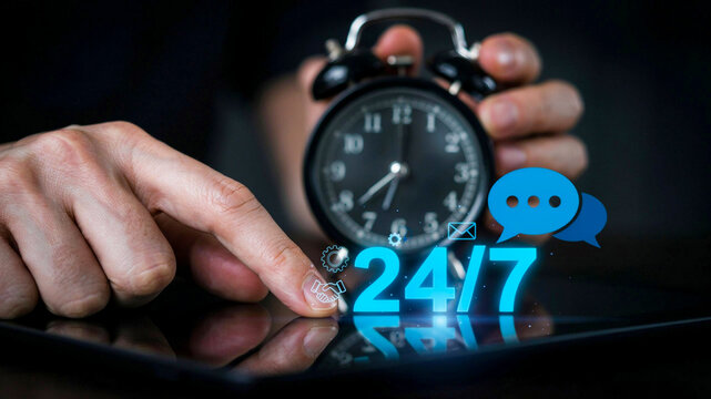 Hand reaching for a digital tablet featuring a twenty four seven availability concept icon against an old fashioned analog alarm clock background