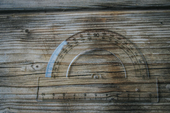 A transparent plastic protractor with an integrated ruler rests on worn wooden boards, displaying angle and centimeter markings.