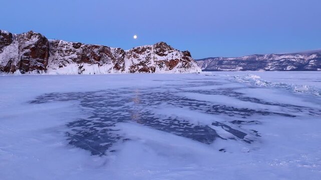 View of the frozen surface of Lake Baikal with snow-covered rocky cliffs and a full moon in the twilight sky in Khuzhir, Irkutsk Oblast, Russia.