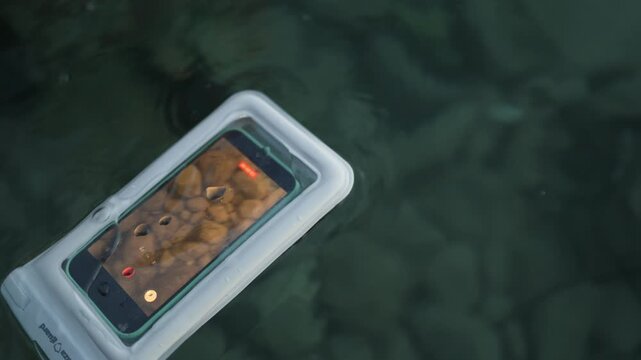 Close up hands hold a smartphone in waterproof case while filming underwater in clear sea showing mobile technology used safely for creative vacation contents and travels