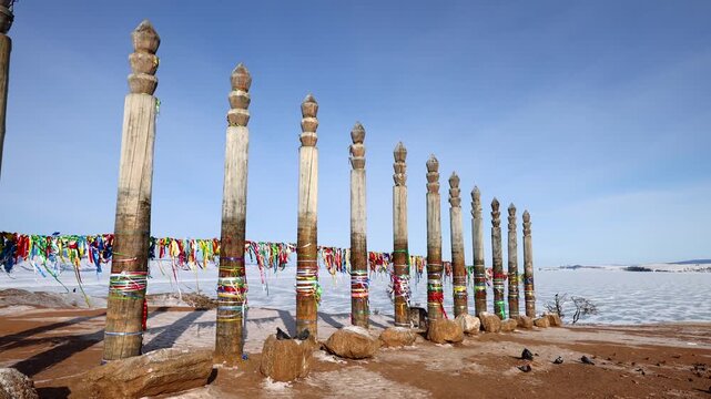 View of ritual wooden poles adorned with colorful ribbons at Lake Baikal, set against a vast frozen landscape under a clear blue sky in Khuzhir, Irkutsk Oblast, Russia.
