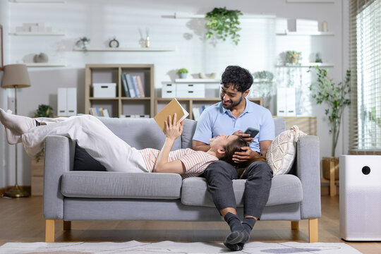 Relaxing couple concept. woman reads a book with her head on man lap as he uses smartphone on sofa at home. Couple, relaxing, lifestyle, reading, smartphone