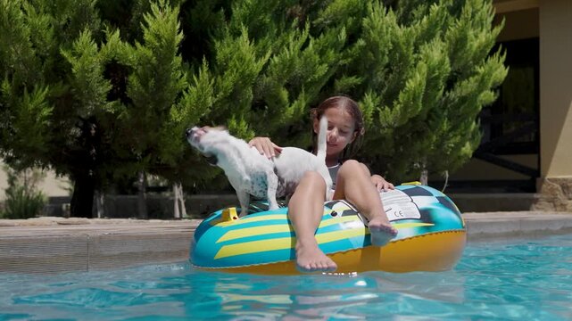 Happy young girl floating on a colorful inflatable tube ring with her pet Jack Russell terrier in an outdoor pool on a sunny summer day