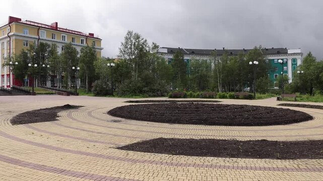 View of a paved circular plaza with dark soil beds and surrounding trees in Kirovsk, Murmansk Oblast, Russia.