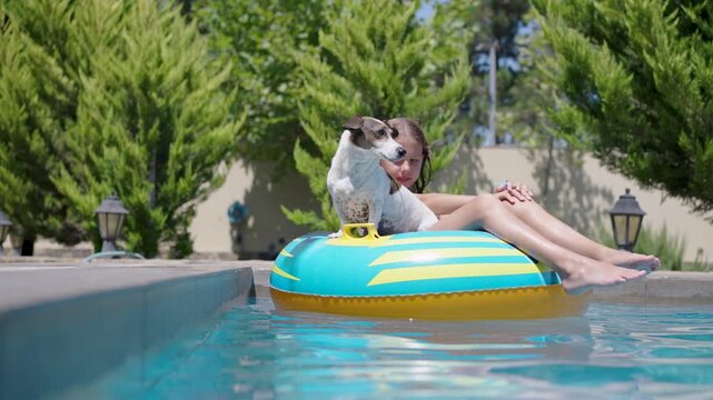 Happy young girl floating on a colorful inflatable tube ring with her pet Jack Russell terrier in an outdoor pool on a sunny summer day