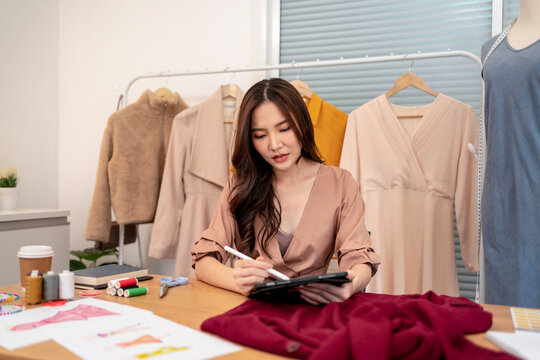 Young asian fashion designer sketching dress design on digital tablet at worktable