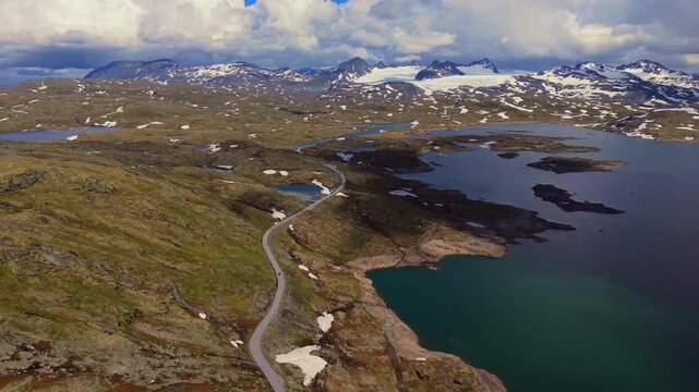 Aerial view of a winding mountain road crossing the rugged highlands of Jotunheimen National Park, Norway, surrounded by alpine lakes, patches of snow and dramatic snow-covered peaks