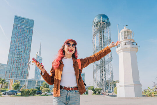Young woman with pink hair smiling, taking a selfie on a smartphone while visiting Batumi, Georgia with city landmarks