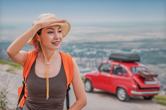 Asian woman hiker looking at the horizon next to her vintage red car parked on the mountain road