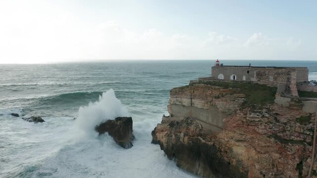 Aerial view of powerful Atlantic ocean waves crashing against rocky cliffs and lighthouse in Nazare Portugal.