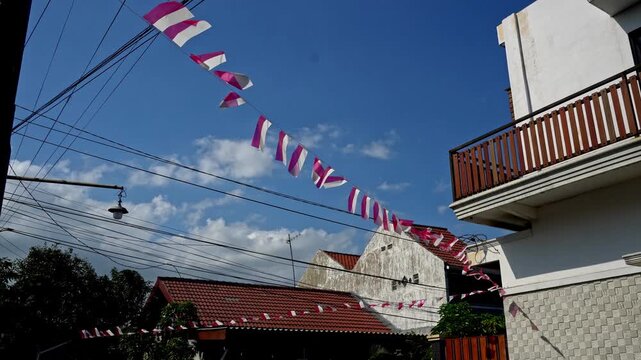 Red and white Indonesian national flags hanging across a residential street and building facade, celebrating independence day under a bright sunny blue sky.