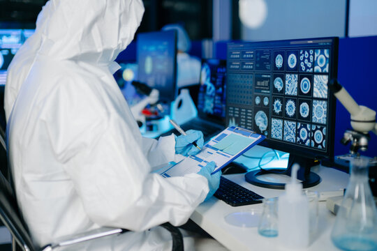Medical researcher in protective suit reviewing and recording data from multi screen digital displays inside research laboratory.