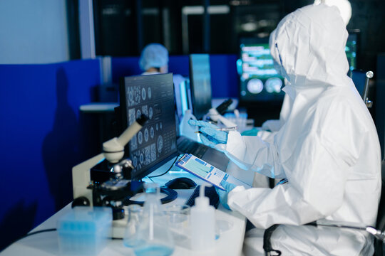 Medical researcher in protective suit reviewing and recording data from multi screen digital displays inside research laboratory.