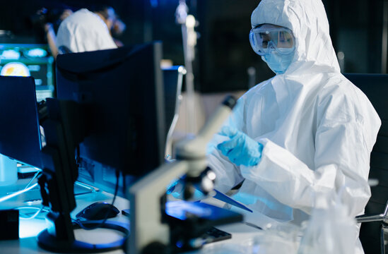 Medical researcher in protective suit reviewing and recording data from multi screen digital displays inside research laboratory.