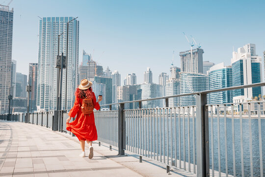 Woman wearing red dress and hat walking along modern urban waterfront promenade with skyscrapers on a sunny day in Dubai