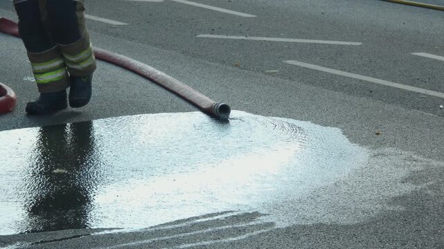 Water flowing from disconnected fire hose across wet asphalt after firefighting operation. Red emergency hose on roadway beside traffic cone. Fire suppression equipment and water runoff on urban