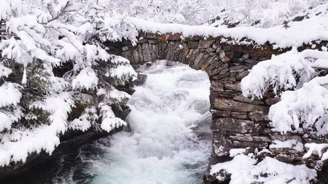View of an old stone bridge spanning a rushing river surrounded by snow-covered trees and rocks in Valldal, More og Romsdal, Norway.