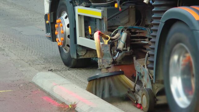 Street sweeper cleaning road surface beside curb before asphalt resurfacing work. Rotating industrial brush removing dirt and debris from roadway. Municipal road maintenance equipment preparing