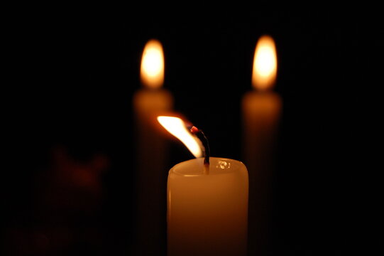 Close-up of a burning candle with a bright glowing flame reflected in a dark surface, isolated against a black background. Atmospheric low-key lighting creates a warm, dramatic and mysterious mood.