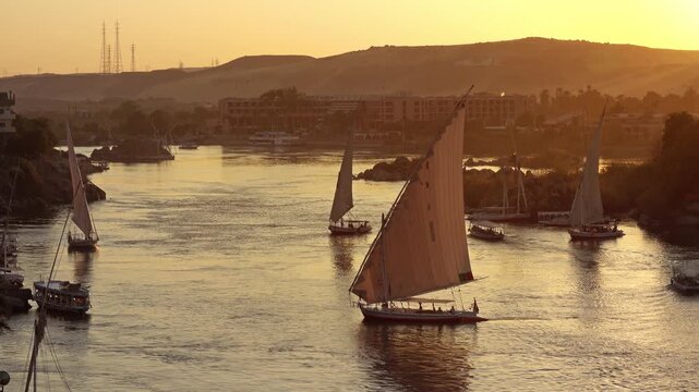 View of traditional felucca sailboats gliding on the Nile River during a golden sunset in Aswan, Aswan Governorate, Egypt.
