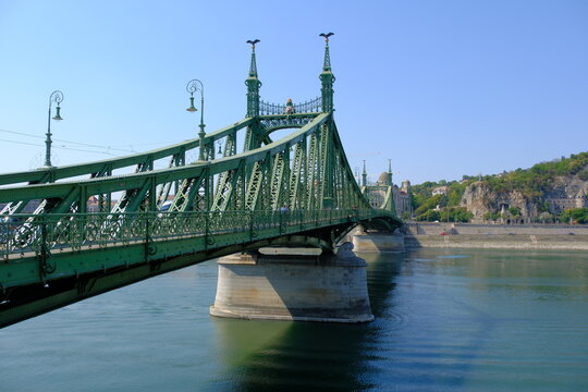 Wide shot of the historic green steel Liberty Bridge (Szabads&aacute;g h&iacute;d) spanning the Danube River in Budapest, featuring its ornate ironwork, turul bird statues atop the spires, and Gell&eacute;rt Hill