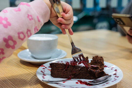 Child Reaching With Fork to Chocolate Cake Slice Beside Coffee Cup on Cafe Table