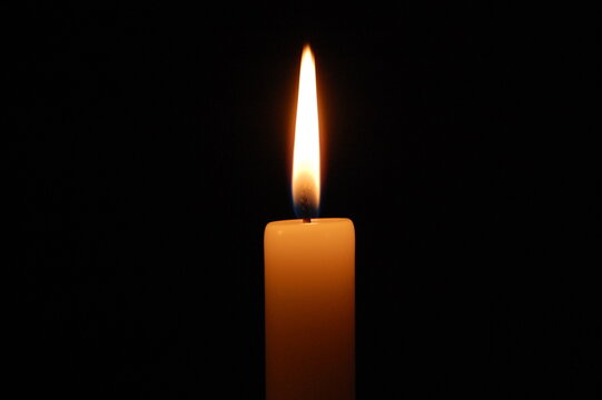 Close-up of a burning candle with a bright glowing flame reflected in a dark surface, isolated against a black background. Atmospheric low-key lighting creates a warm, dramatic and mysterious mood.